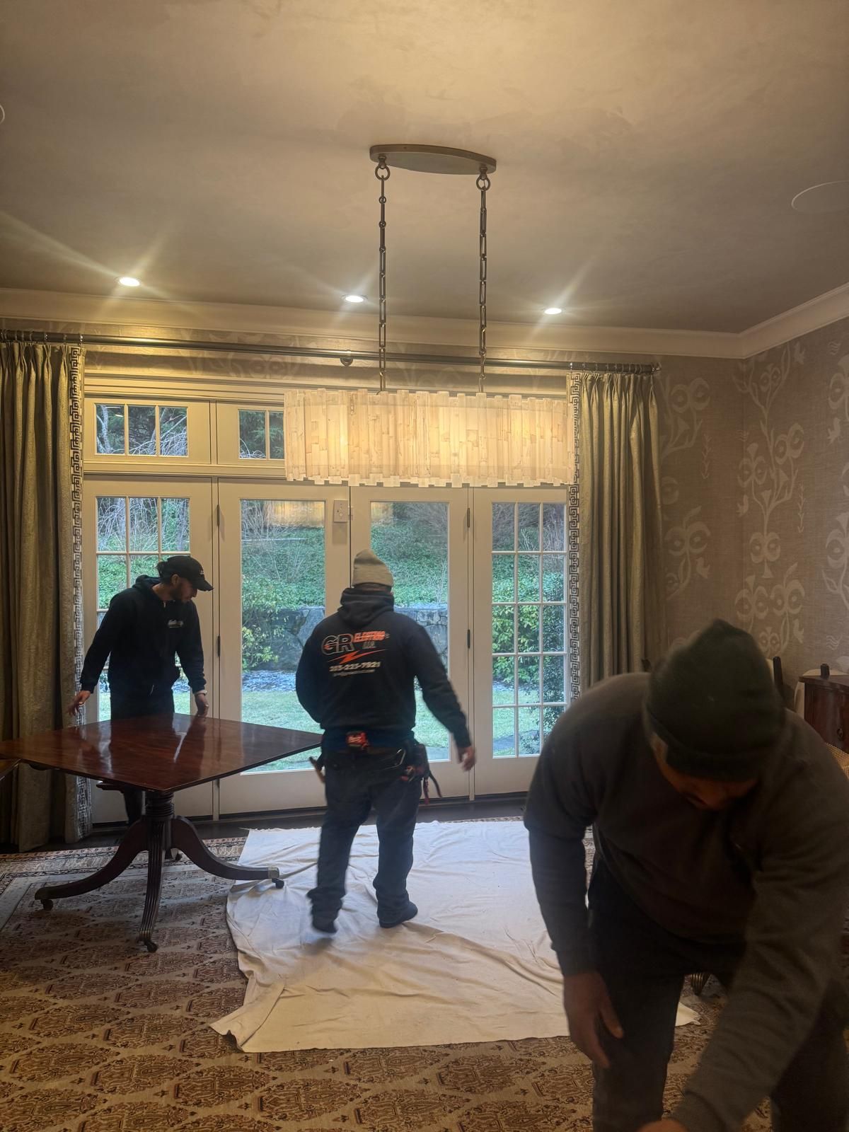 Three people setting up a round table on a rug near glass doors. A light fixture hangs overhead.