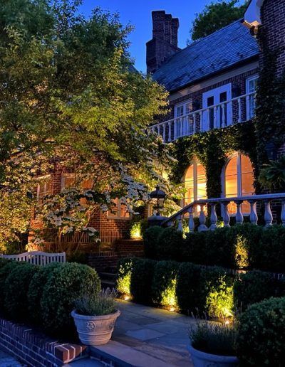 Night-lit brick house with ivy, balcony, and manicured hedges. Pathway and potted plants are in the foreground.