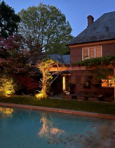Poolside view of a brick house at dusk, with lights illuminating the surrounding foliage and reflecting in the water.