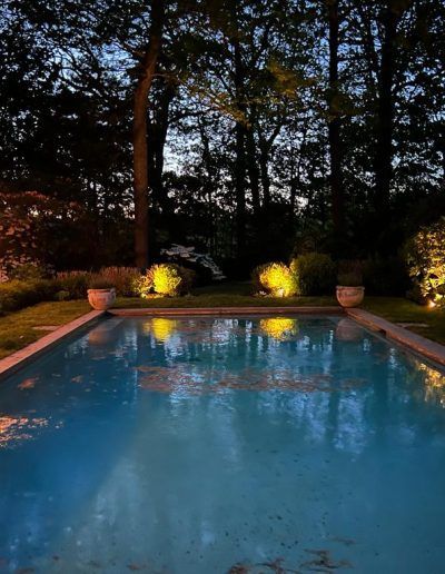 Swimming pool at dusk, illuminated by landscape lighting in a backyard, surrounded by trees.