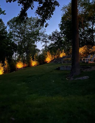 Evening view of a grassy hill illuminated by orange lights. Trees line the top of the hill, and a blue sky is visible.