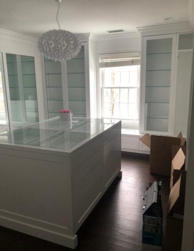 White walk-in closet with island, glass-front cabinets, and window. Dark wood floor.