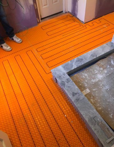 Orange radiant floor heating system installed on a floor, with a person's legs and shoes visible.