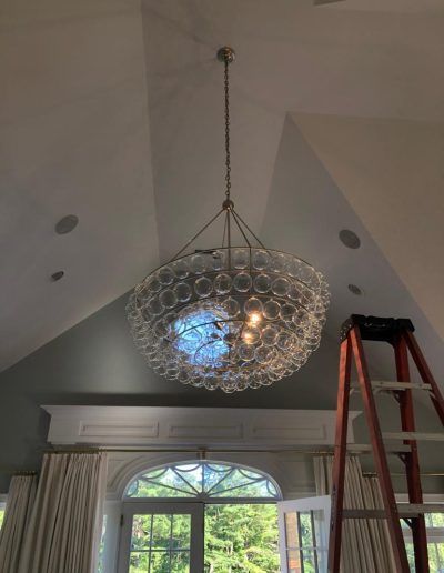 A large, round glass chandelier hangs from a high ceiling; a ladder rests nearby.