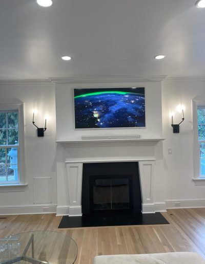 Living room with fireplace, TV, and sconces. White walls, wood floor, and two windows.