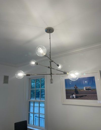 Modern chandelier with five globe lightbulbs hangs from a white ceiling, near a window and framed artwork.