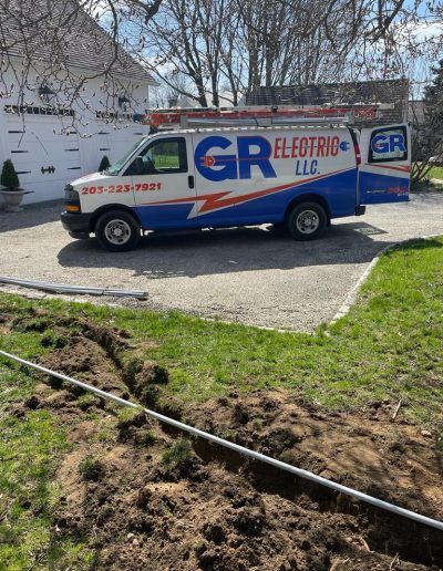 White and blue GR Electric LLC van parked next to a trench in the grass. A ladder is on top.