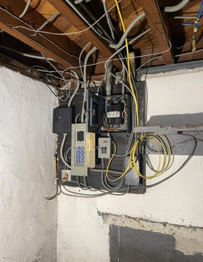 Electrical panel with various wires, boxes, and a control unit on a wall in a basement setting.
