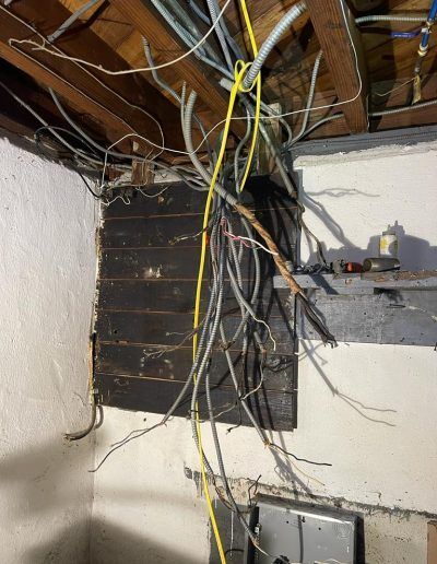 Exposed electrical wires hang from a ceiling in a room with damaged walls and a wooden beam.