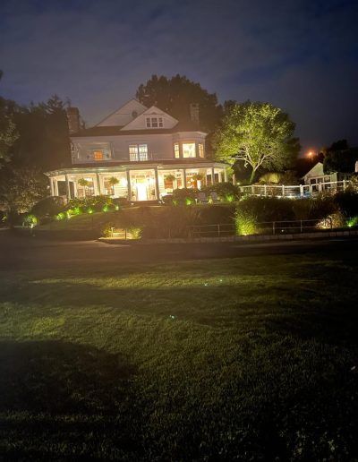 White house at night, lit by exterior lights, with a long porch and a green lawn.