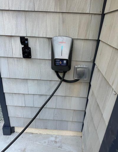 Electric vehicle charger mounted on gray shingle siding with black cable and outlet.