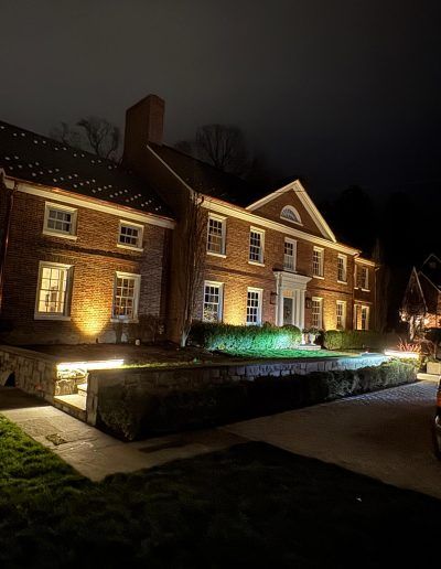 A brick house illuminated at night. Features include a white trim, windows, and manicured landscaping.