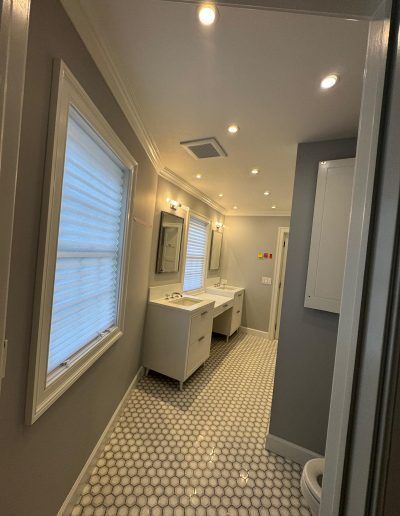 Bathroom with white cabinets, mirrors, and honeycomb tile floor. Gray walls, recessed lighting, and a window with blinds.