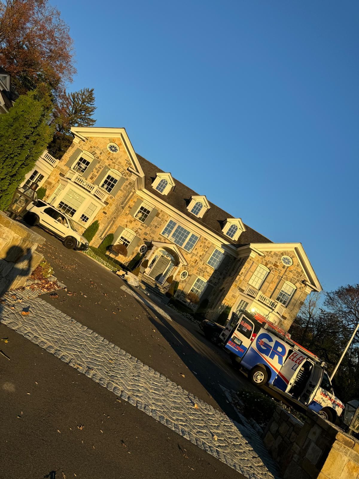 Stone house with service van parked on driveway. Blue sky, fall foliage visible.