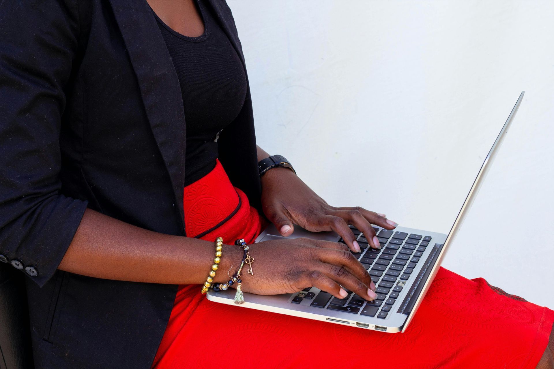 Black woman in blazer types on laptop outdoors. Red skirt, black top, white background.