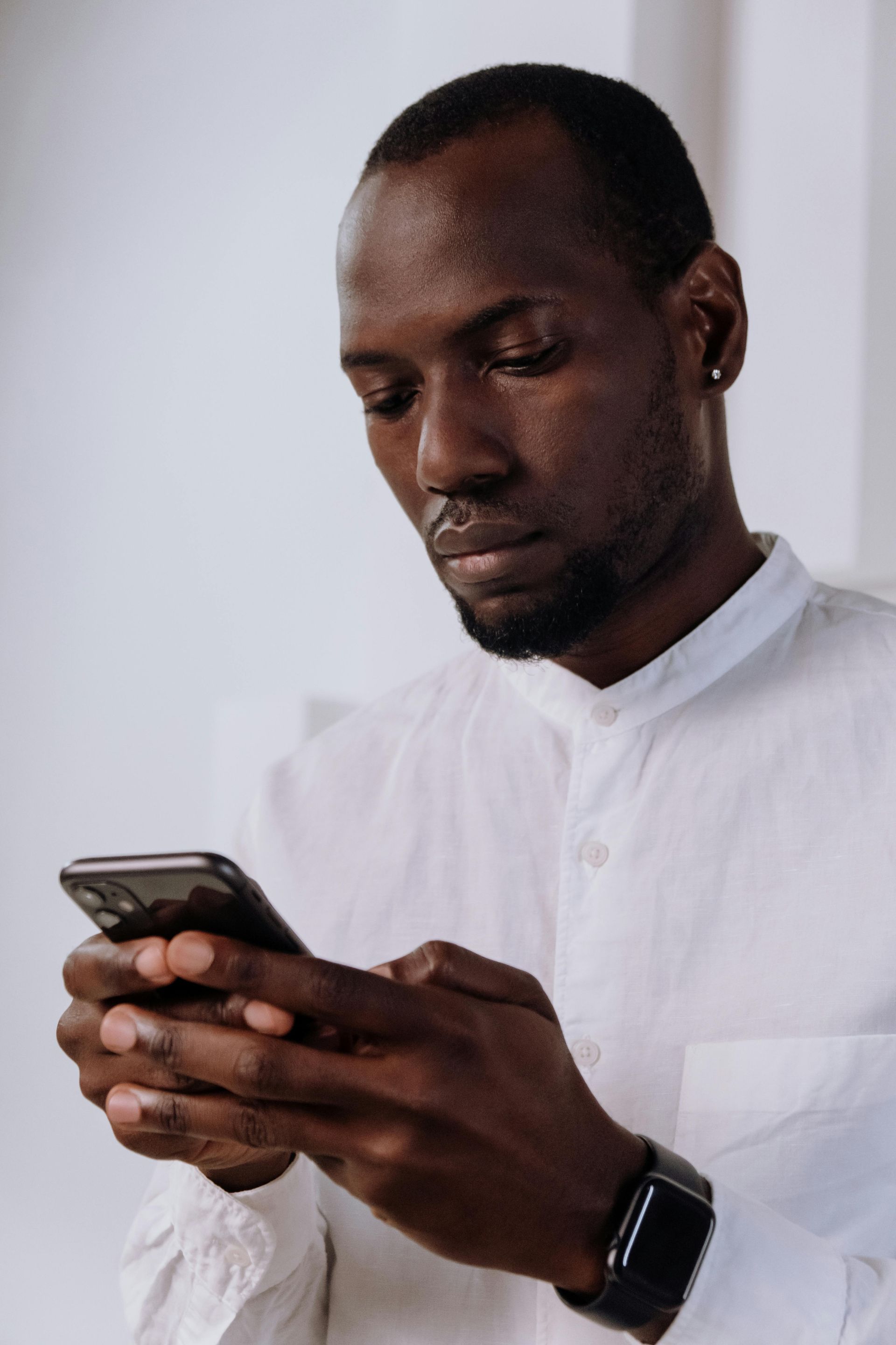 Man in white shirt using a smartphone, focused expression. Indoors, neutral background.