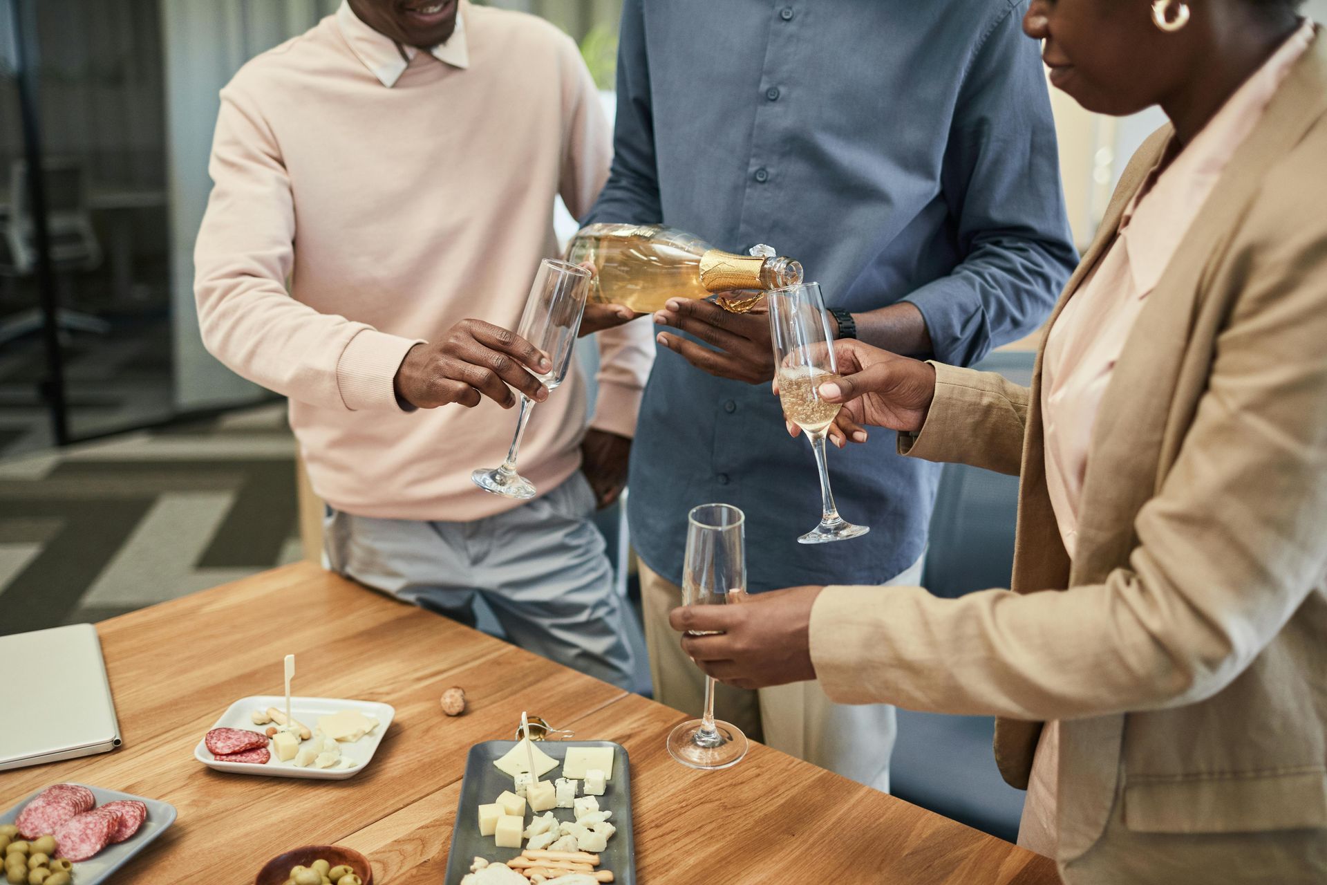 Three people celebrating, pouring champagne. Wooden table with cheese, meat.