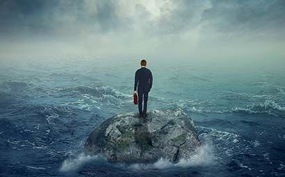 Man standing on a small rock in a stormy sea, holding a briefcase, overcast sky.
