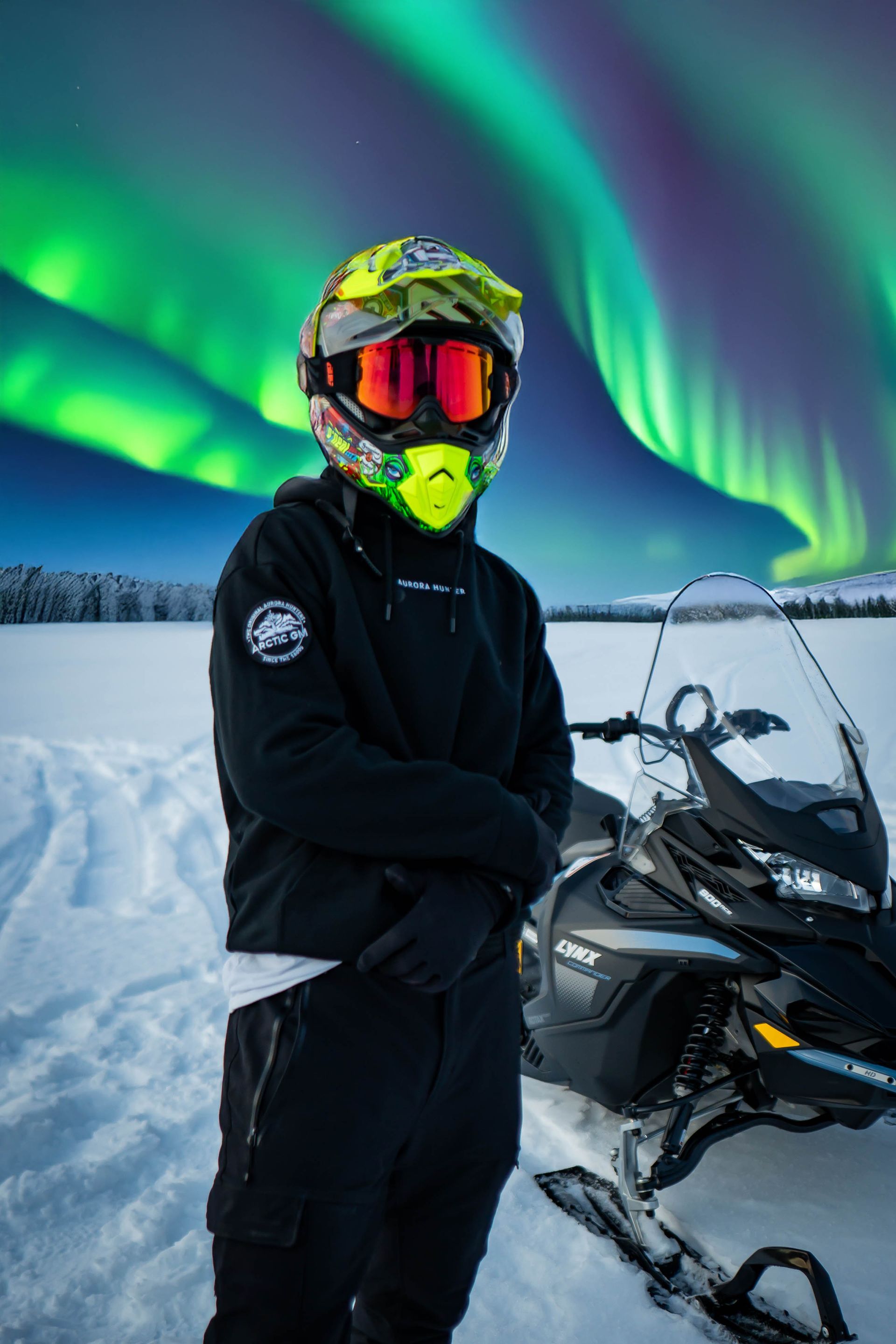 Snowmobiler in Arctic GM hoodie and helmet standing under the Northern Lights