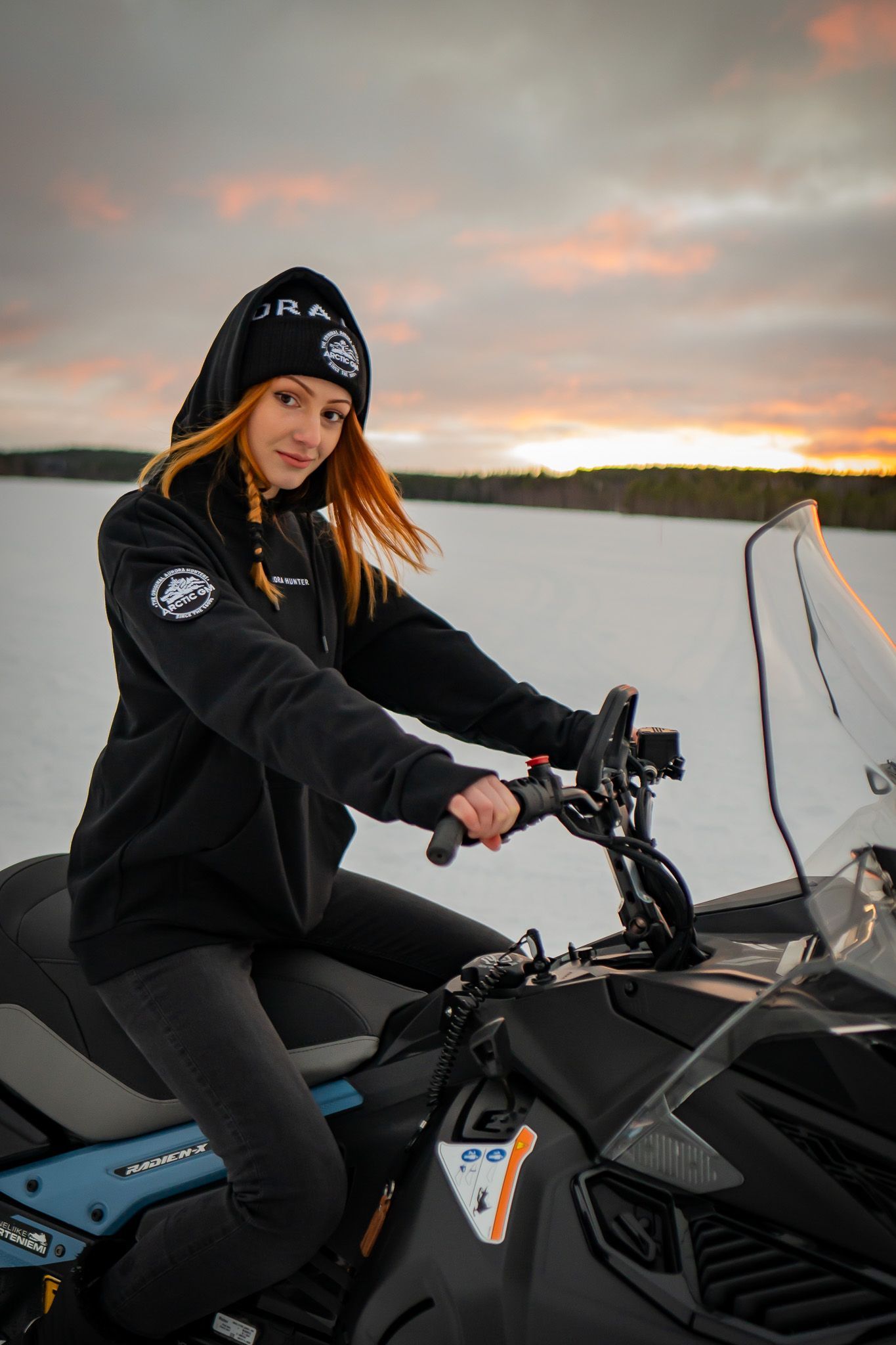 Woman riding snowmobile in Arctic GM hoodie and beanie at sunset in Lapland