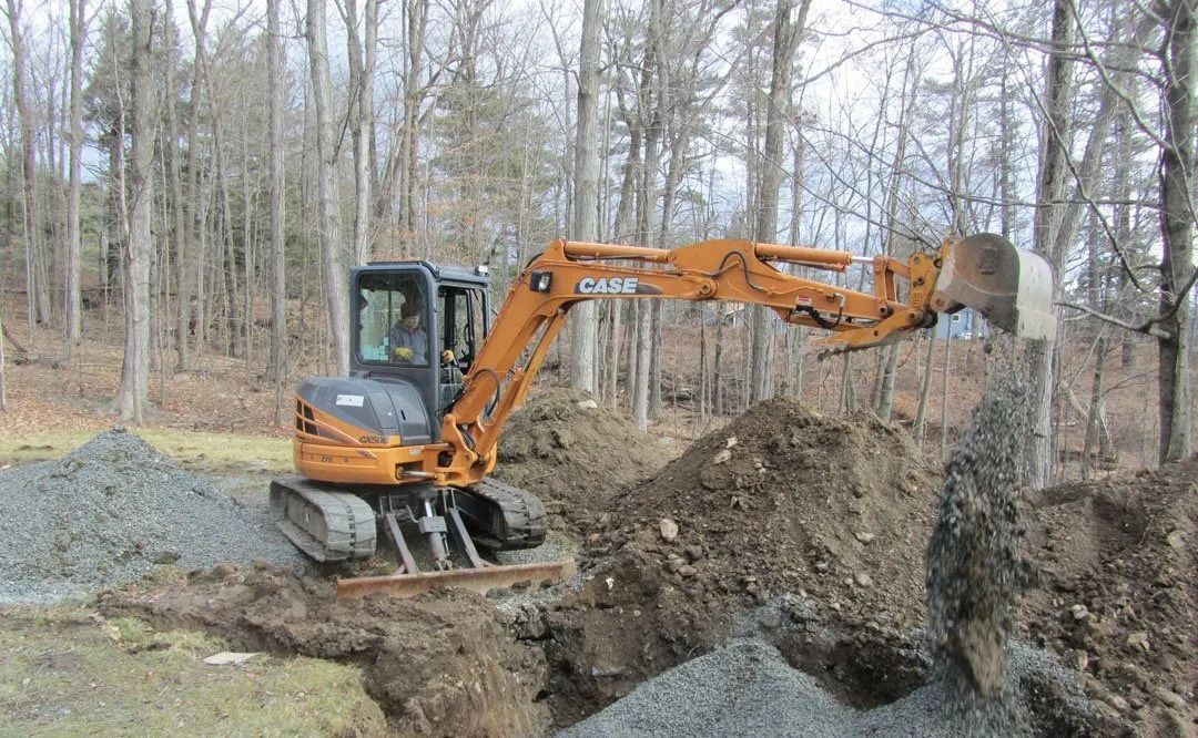 A small excavator is digging a hole in the ground in the woods.