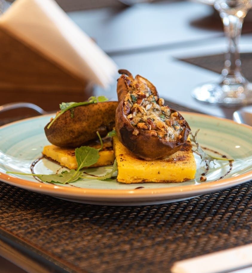 Plato de comida: pera asada con relleno, polenta y hierbas en un plato con borde verde azulado.
