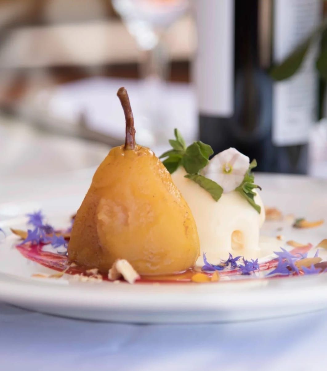 Postre de pera escalfada con helado, almíbar, nueces y flores en un plato blanco.