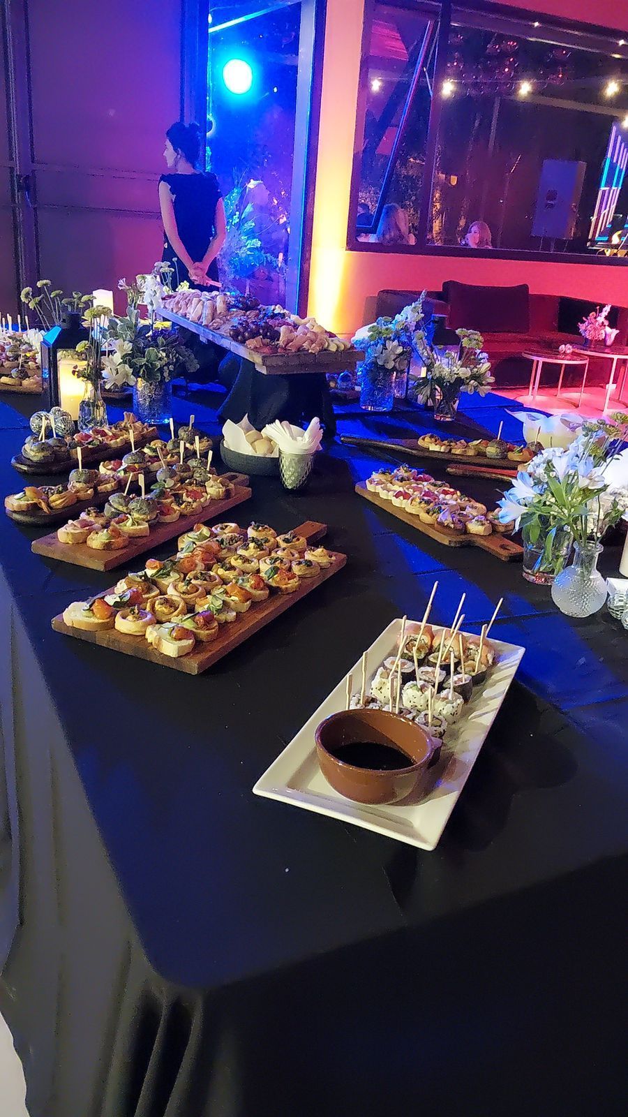 Mesa de buffet con aperitivos sobre tablas de madera y mantel negro. Local iluminado.