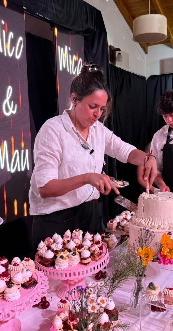 Personas cortando un pastel de varios pisos en un evento con postres. Letrero de neón al fondo.