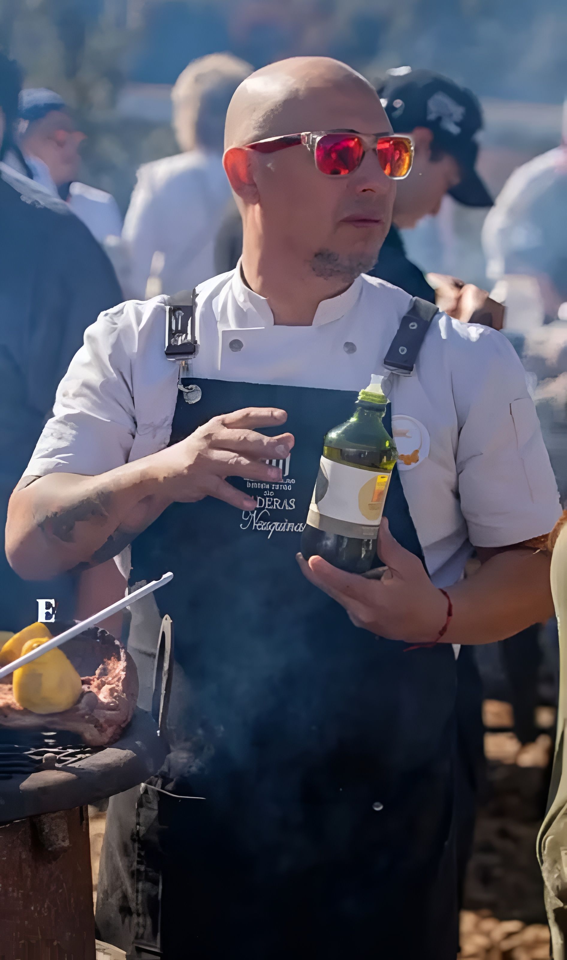 Hombre preparando comida en una cocina al aire libre con fogata. Comida en una mesa, luces de colores en el techo.