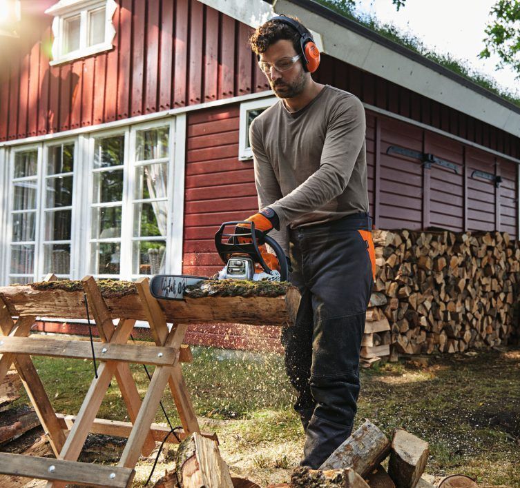 A Man Is Cutting a Log with A Chainsaw in Front of A Red House — Stihl Shop Grafton in Grafton, NSW