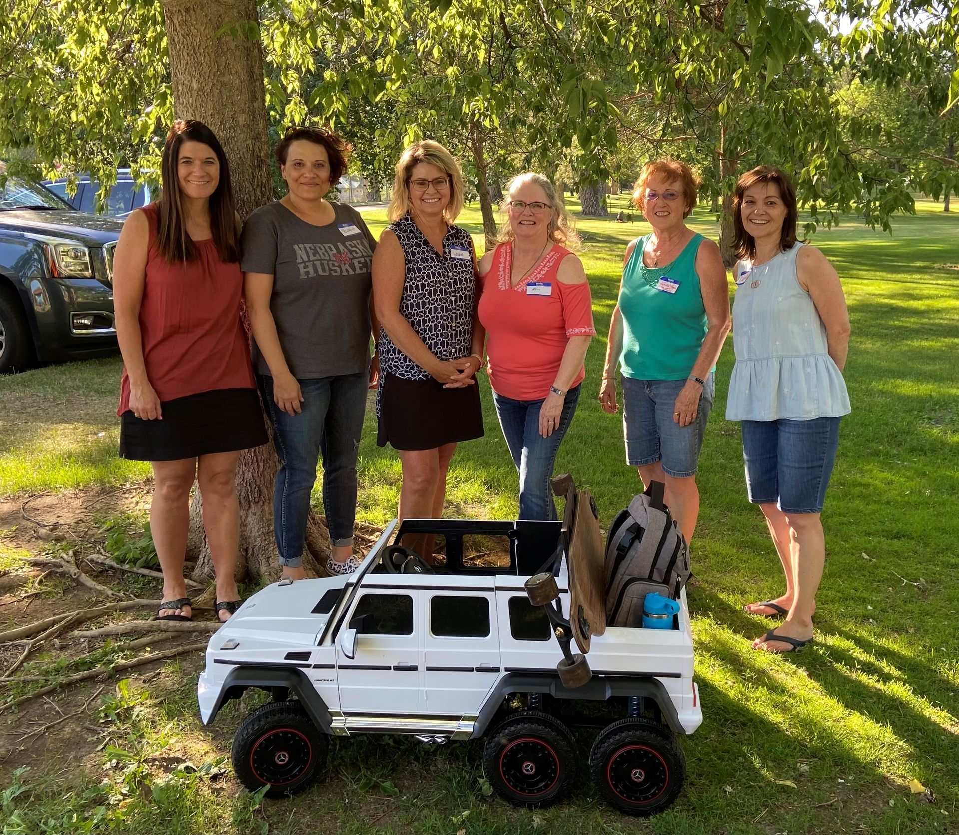 Six women pose near a child's white toy car outdoors.