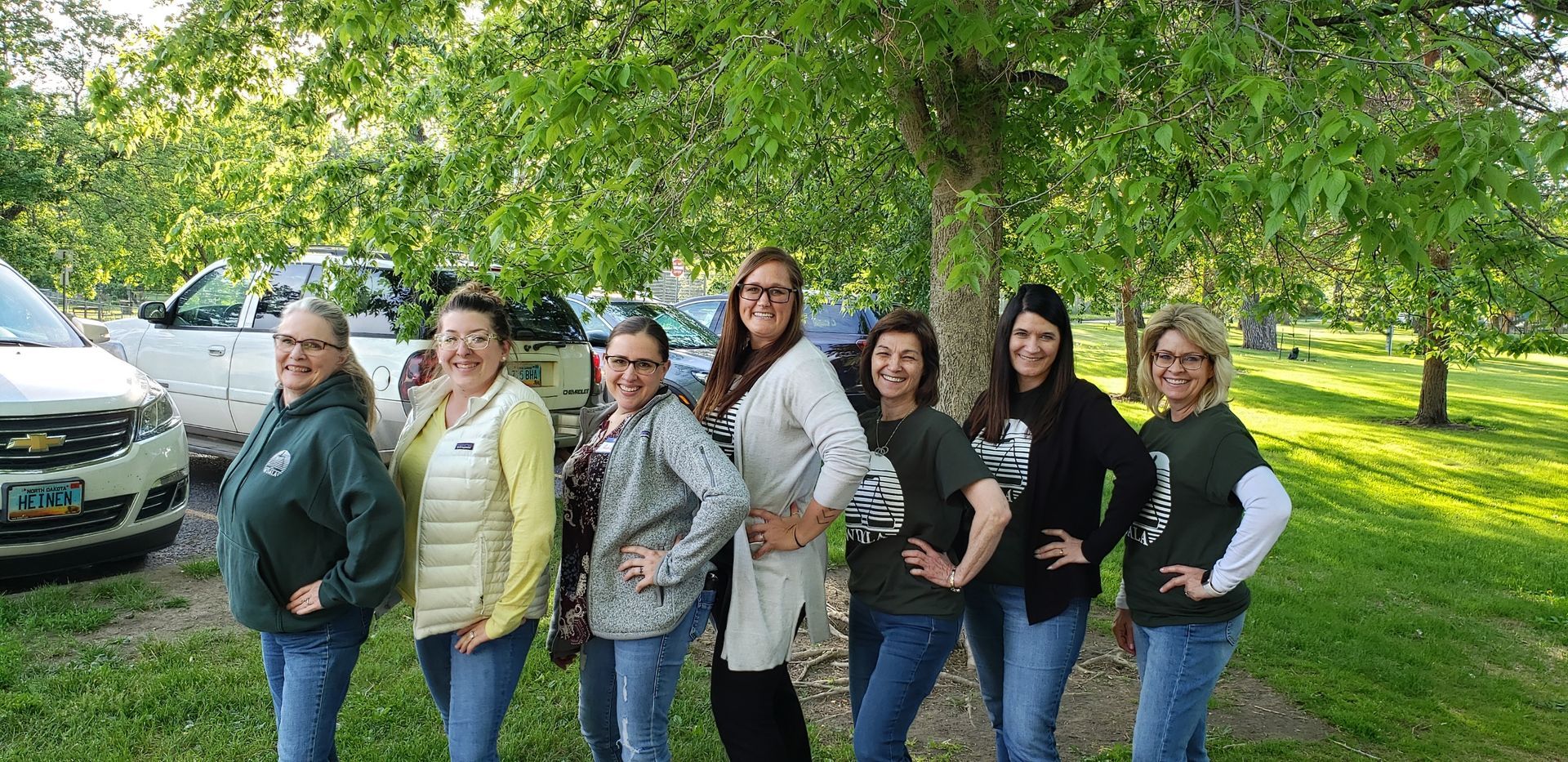 Seven women in jeans pose in front of trees, a park, and vehicles.