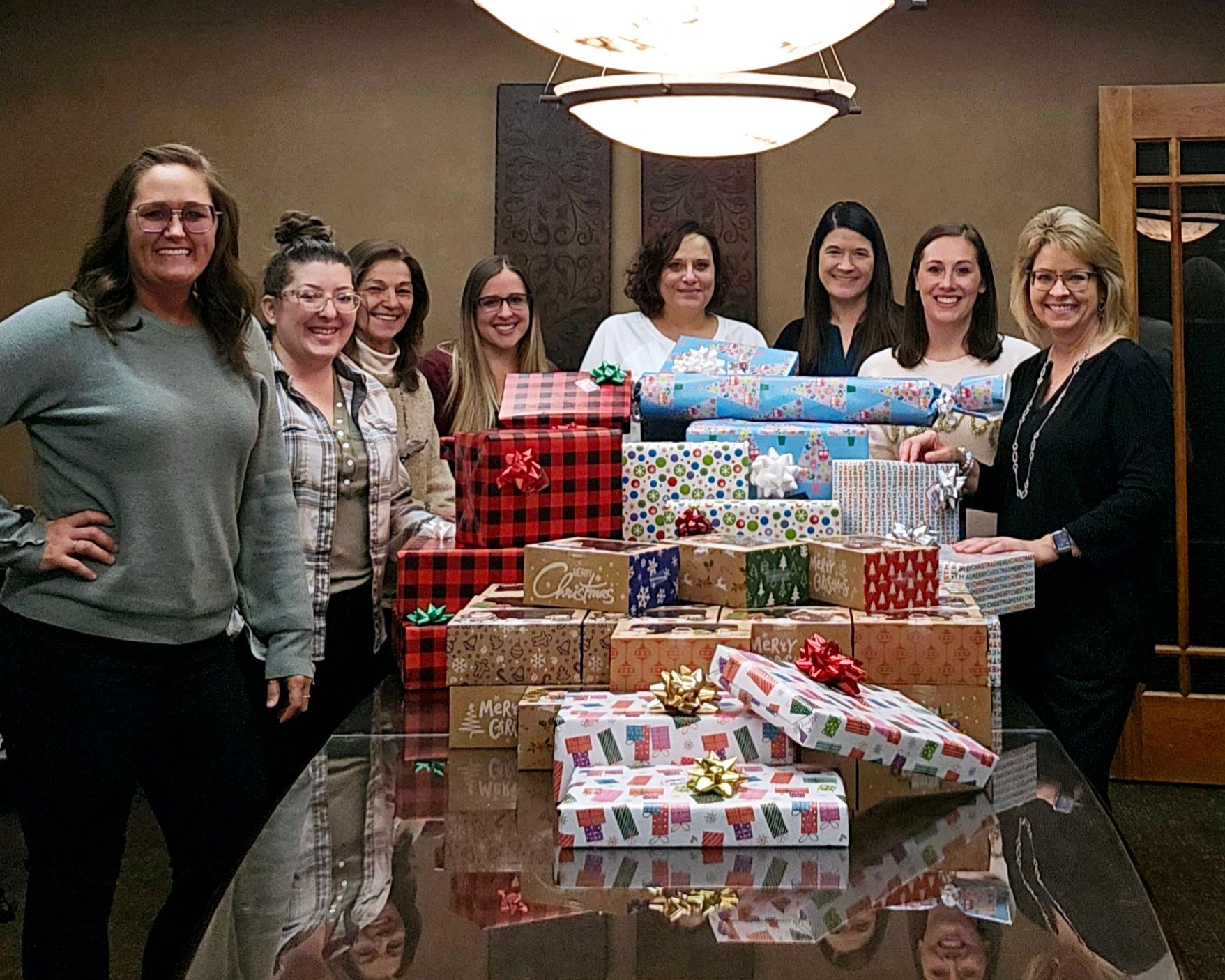 Group of women smiling, gathered around a table laden with wrapped gifts, likely for a holiday.