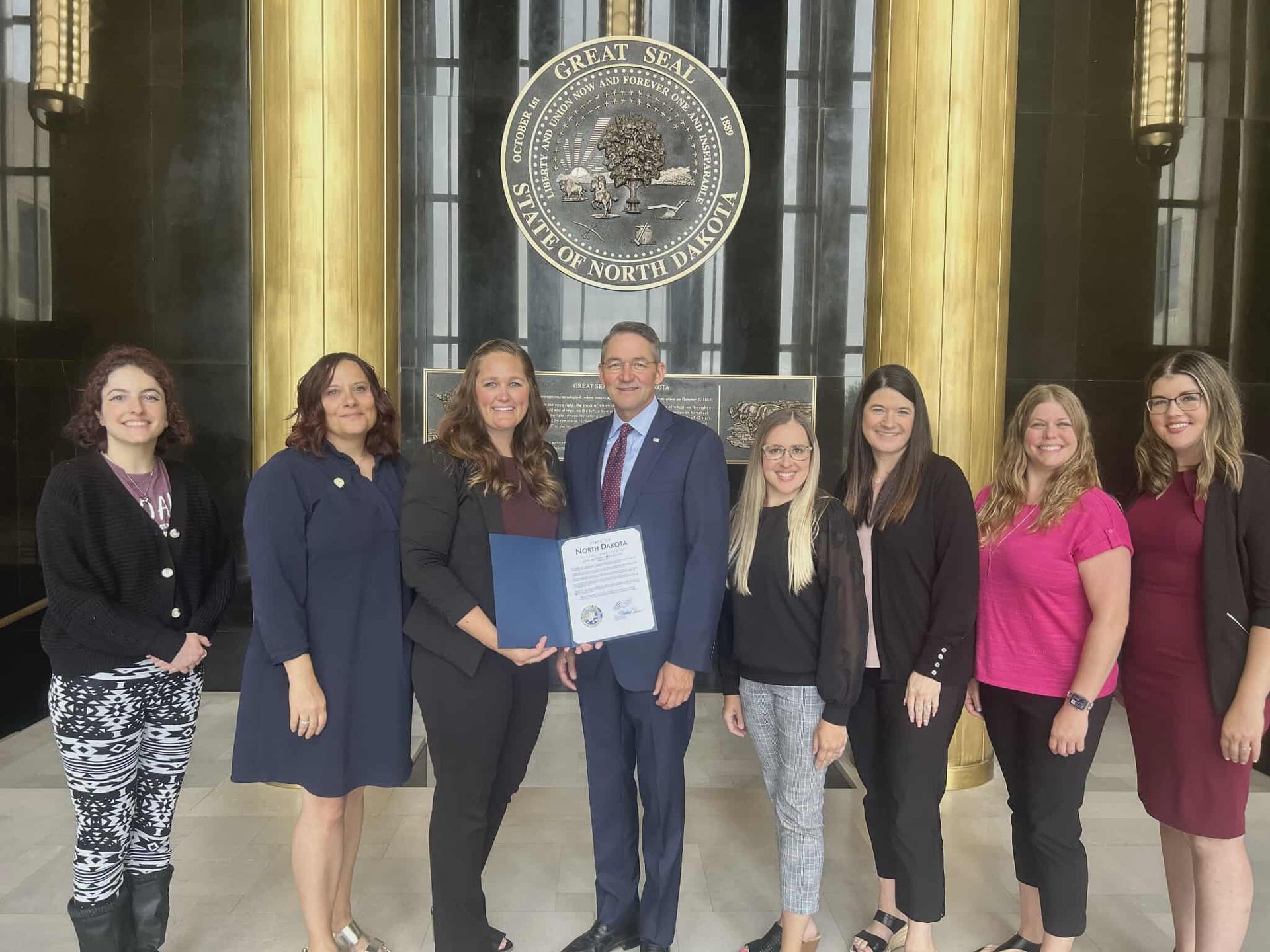 Group of people posing with a man holding a document in front of a large emblem and gold columns.