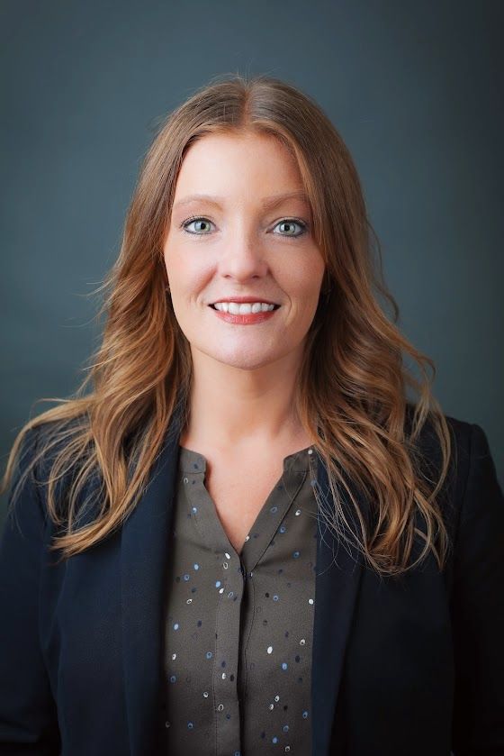 Woman with wavy auburn hair, smiling, wearing a dark blazer and patterned top, against a blue background.