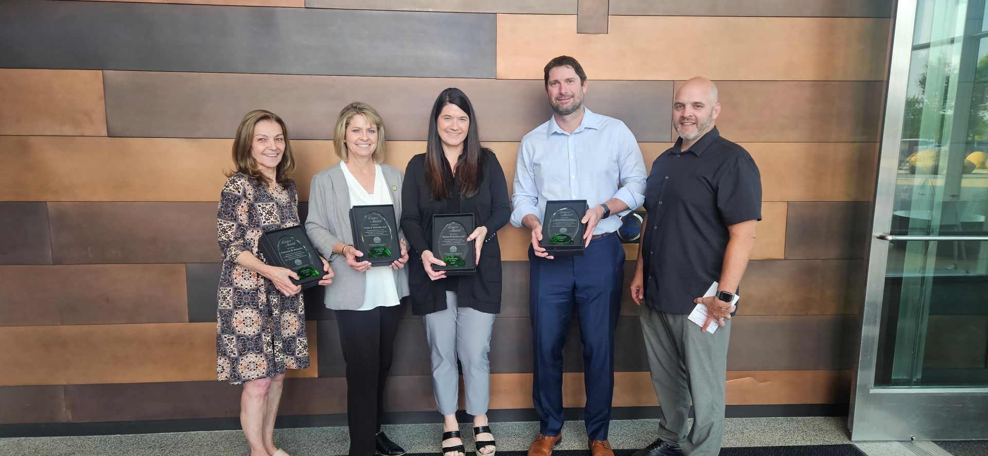 Five people holding awards in front of a wood wall. They stand near a glass door.