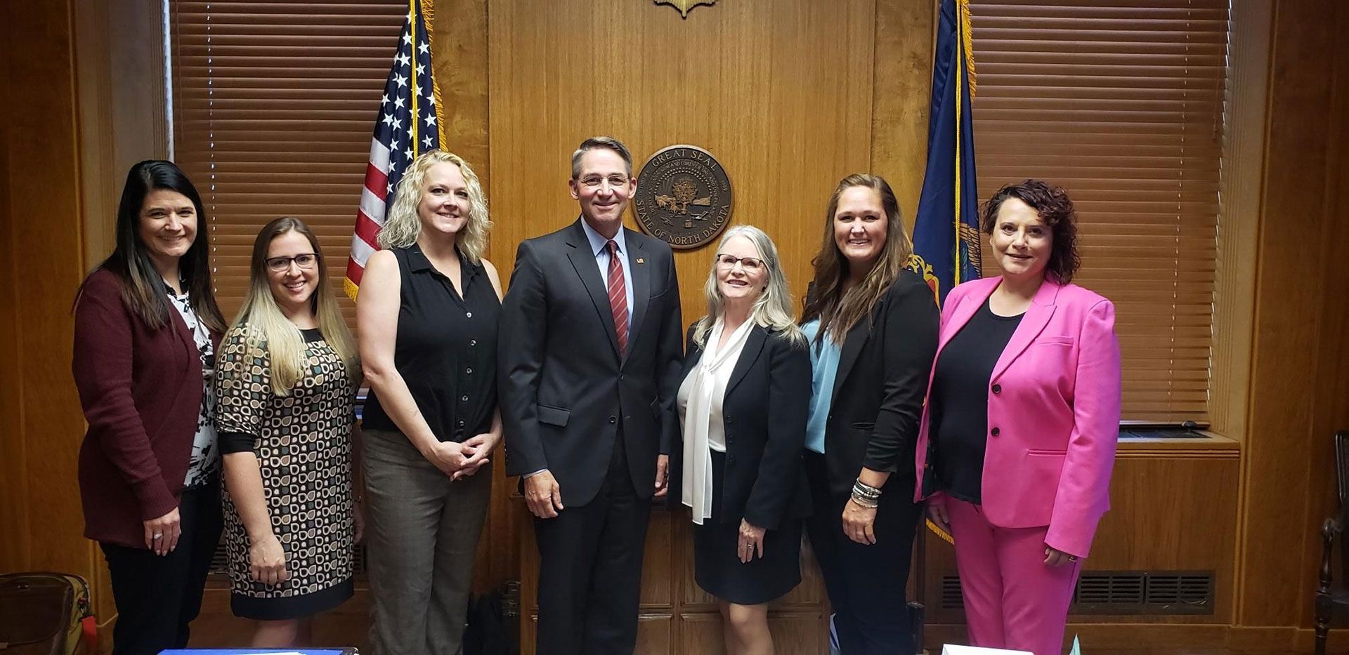 Group of people posing in front of a wooden wall with flags. Seven individuals, some smiling, formal attire.