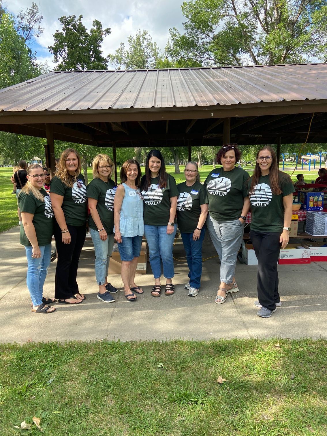 Group of eight people wearing matching green shirts standing under a picnic shelter in a park.