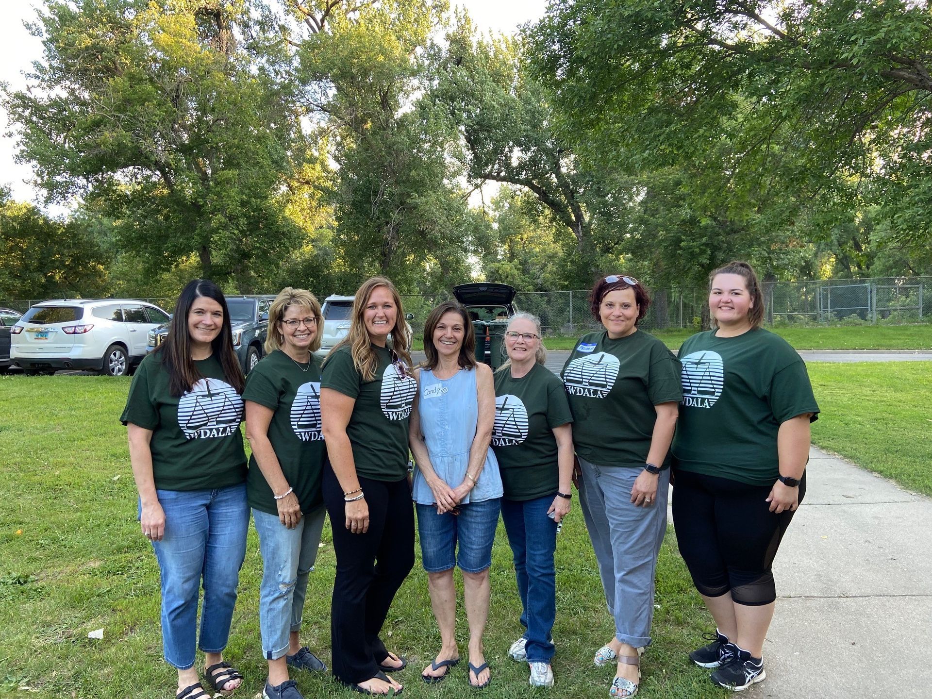 Group of women in green shirts pose outside. Trees and parked cars in the background.