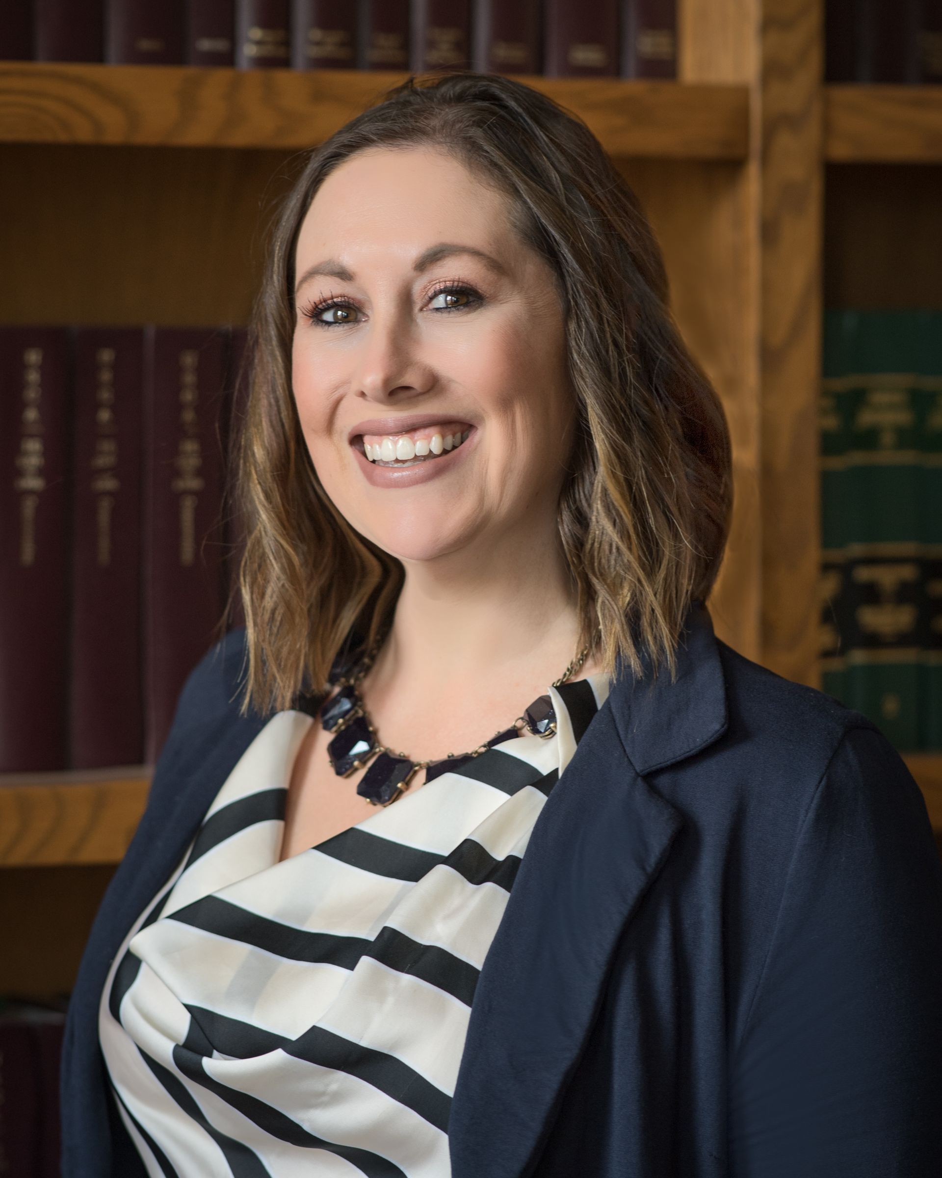 Woman in navy blazer and patterned blouse smiles in front of bookshelves.