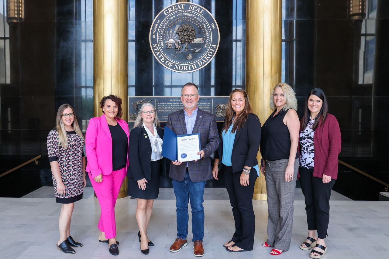Group of seven people in formal attire posing in front of a building with a seal.