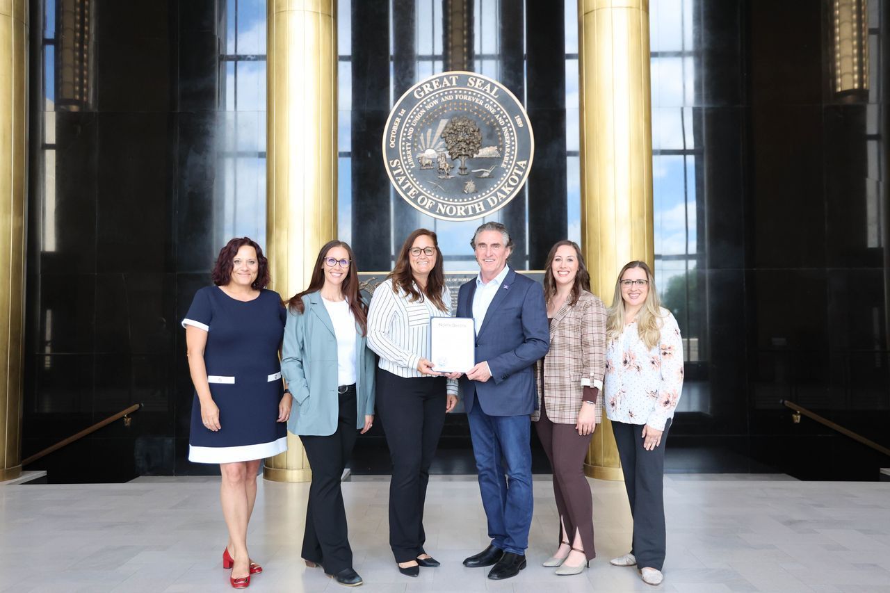 Group of people holding a document; formal setting, large emblem in background.