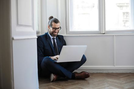 A person with hair tied in a bun, wearing a suit and glasses, sitting cross-legged on a floor, typing on a laptop.
