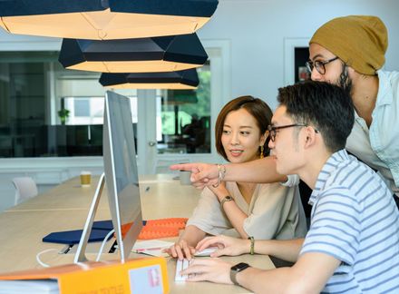 Three people collaborate at an office desk, looking at a computer screen while one points at the display.
