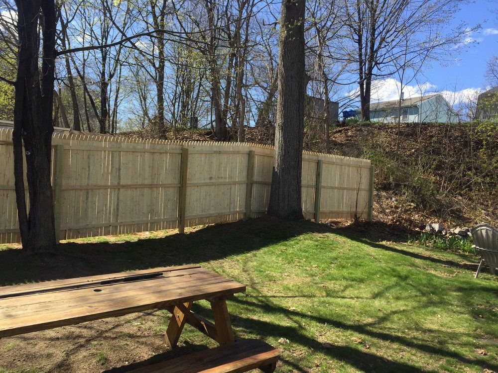 Picnic table in a grassy yard, with a wooden fence and trees in the background under a sunny sky.
