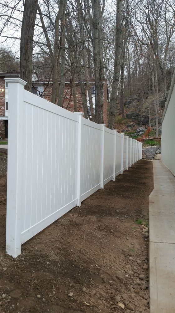White vinyl fence alongside a building and graded dirt, with trees in the background.