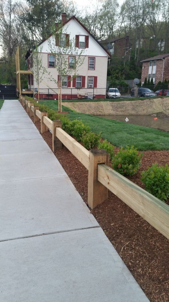 Sidewalk bordered by wooden fence and shrubs, adjacent to a pond and a two-story house.