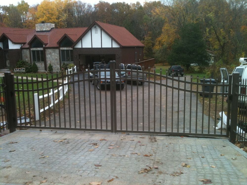 Black metal driveway gate in front of a house. Driveway is brick paved.