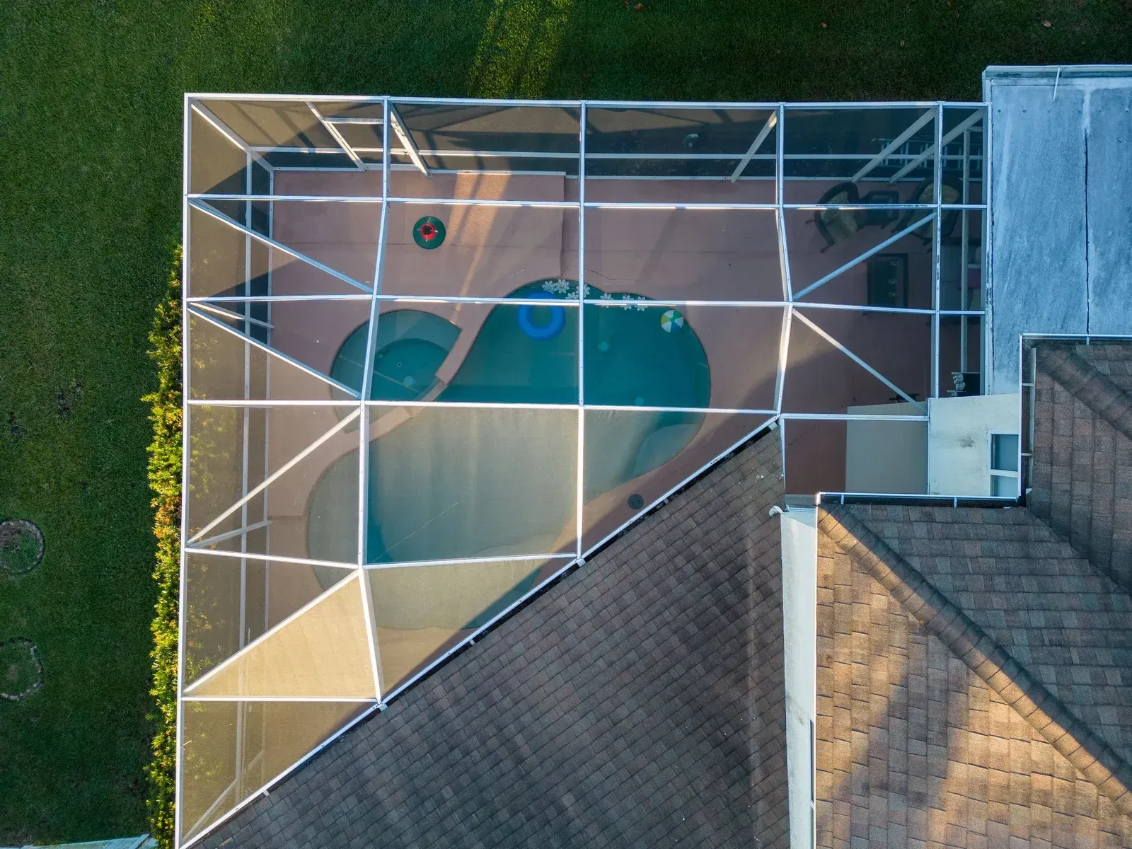 Overhead view of a swimming pool enclosed by a screen. Adjacent to a house with a brown roof.